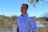 A man wearing a blue button-up shirt stands in front of the river, looking off camera
