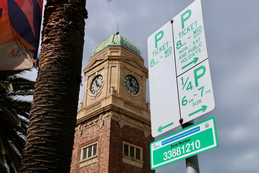 A ticketed car parking sign in front of a clock tower and grey sky.