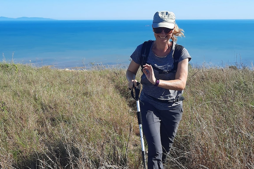 A woman walks through long grass in hiking gear with ocean in background.