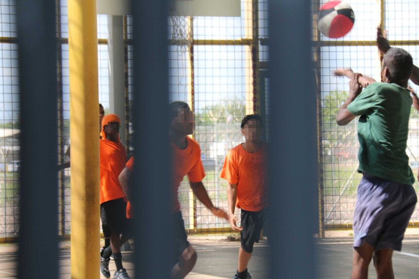 A group of young boys playing basketball in an enclosed space. Most are wearing orange t-shirt and black shorts.