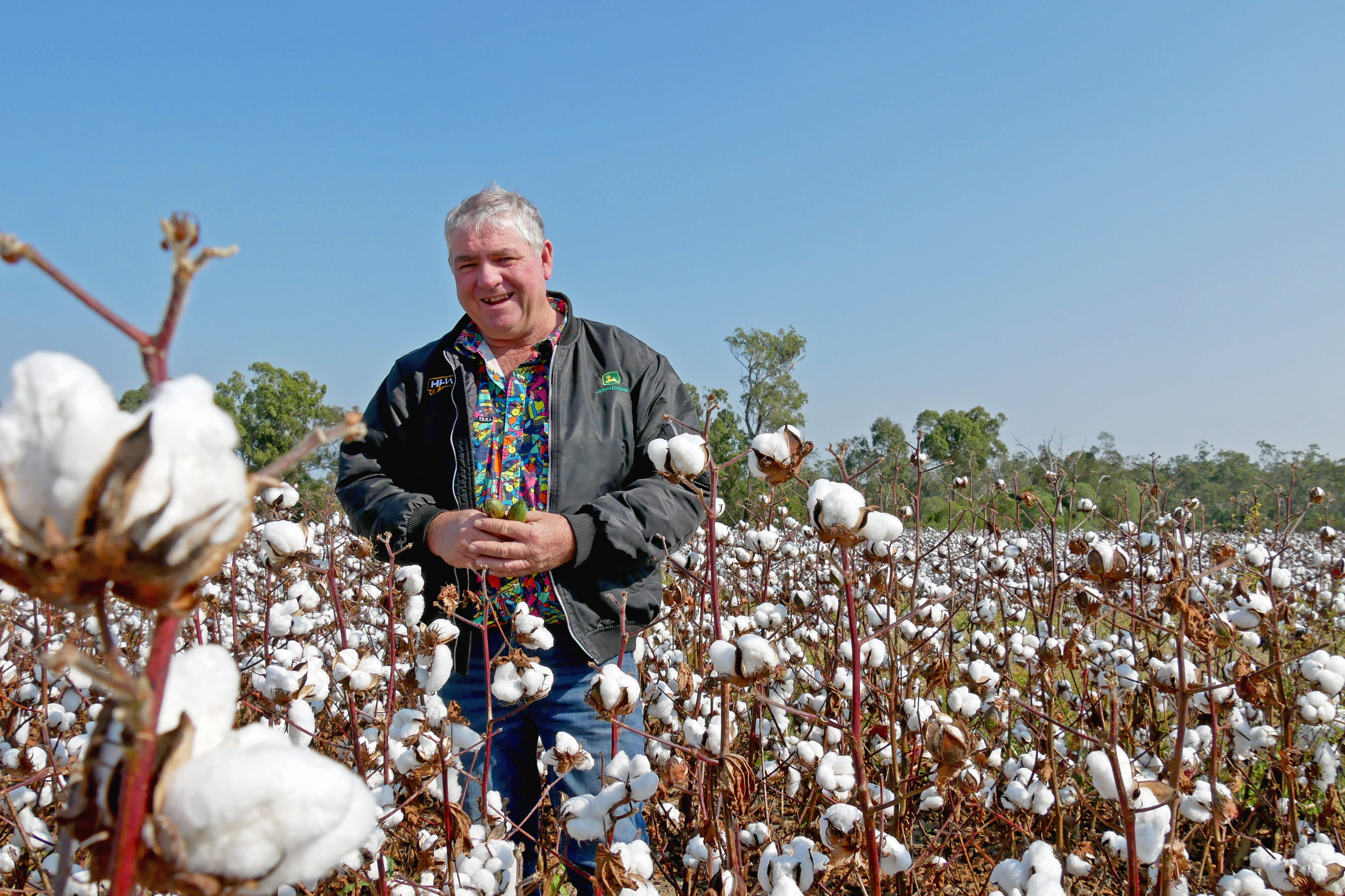 Cotton grower stands in cotton field, smiling looking into camera lense wearing colourful shirt and green jacket
