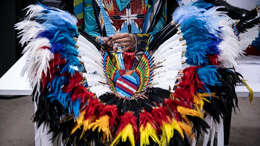Seated on white table, a man ties string of vibrant feathered black, blue, red, green, yellow and white Native American regalia.