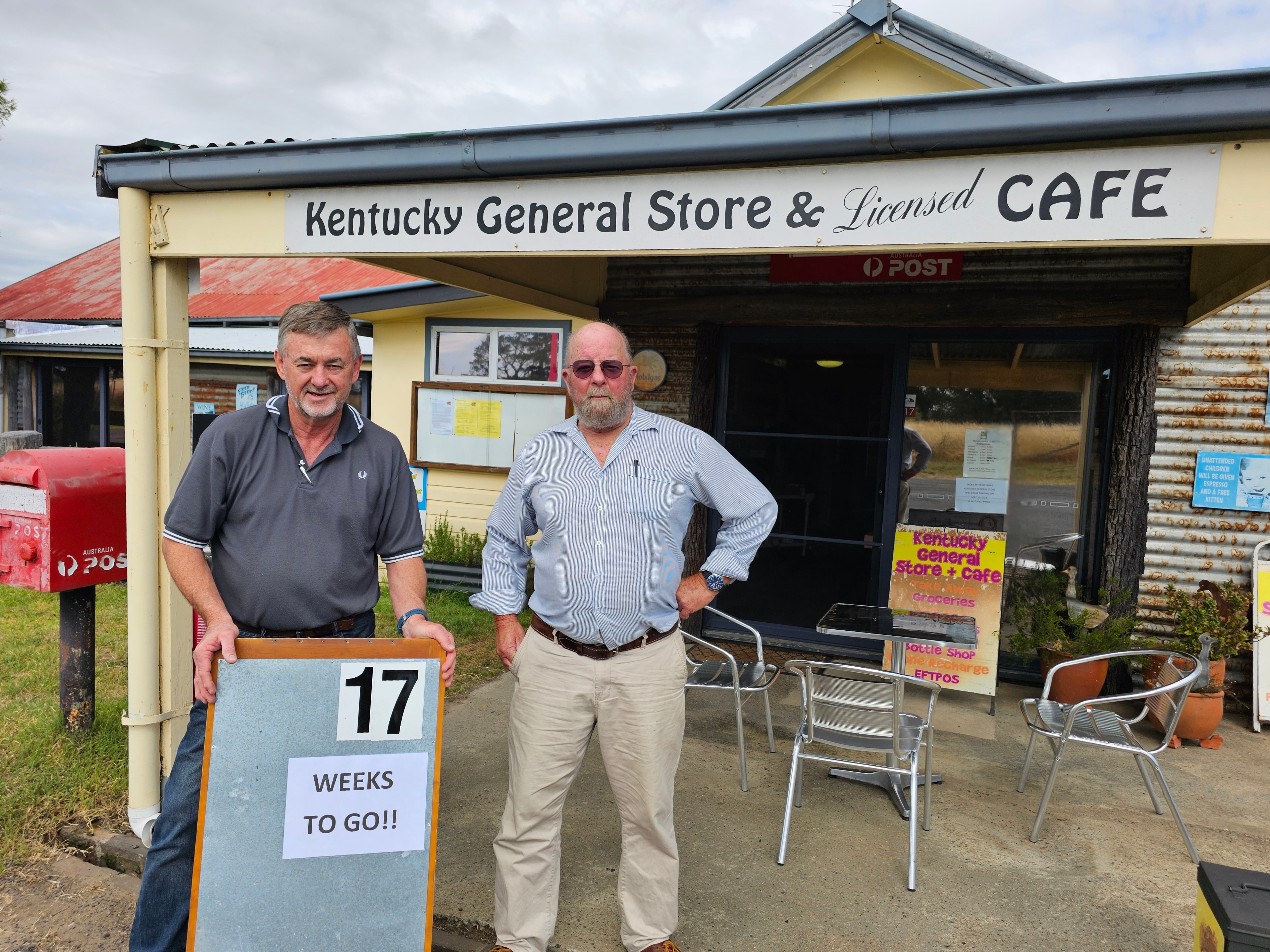 Two men stand in front of th Kentucky General Store with a sign reading "17 Weeks TO GO"
