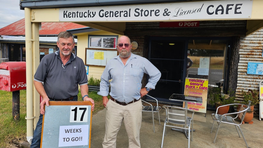Two men stand in front of th Kentucky General Store with a sign reading "17 Weeks TO GO".