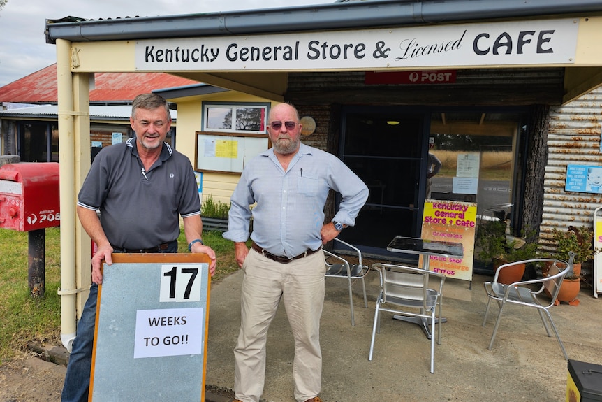 Two men stand in front of th Kentucky General Store with a sign reading "17 Weeks TO GO"