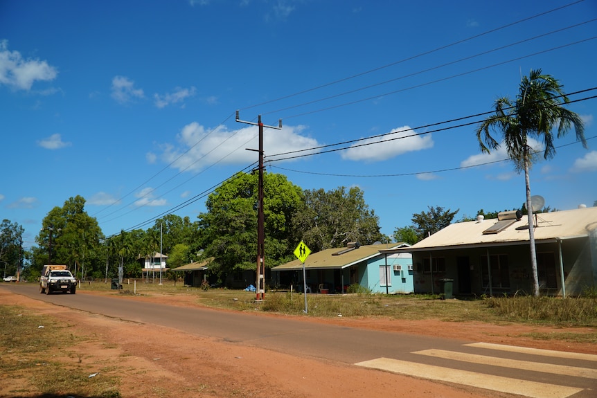 A street view of Pirlangimpi, which shows a police car driving down a road. Housing is basic and there is lots of greenery.