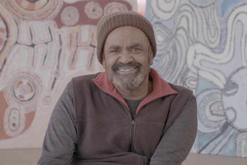 A man wearing a beanie smiles towards camera while sitting on front of two vibrant and large artworks.