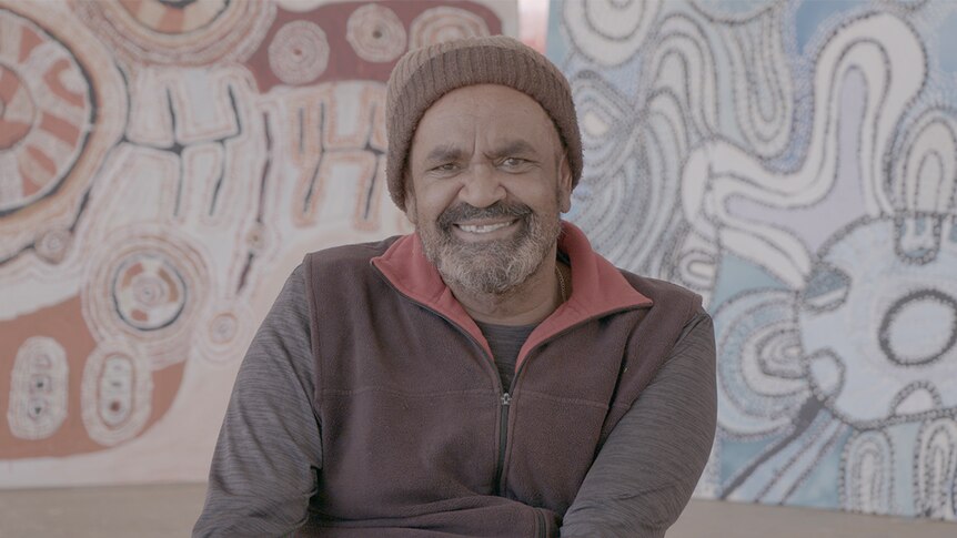 A man wearing a beanie smiles towards camera while sitting on front of two vibrant and large artworks.