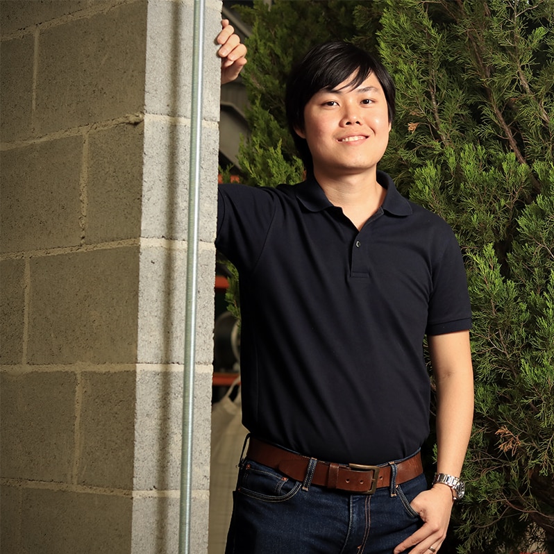 Edward Gan leans against a brick wall in front of some potted trees he will use for testing.