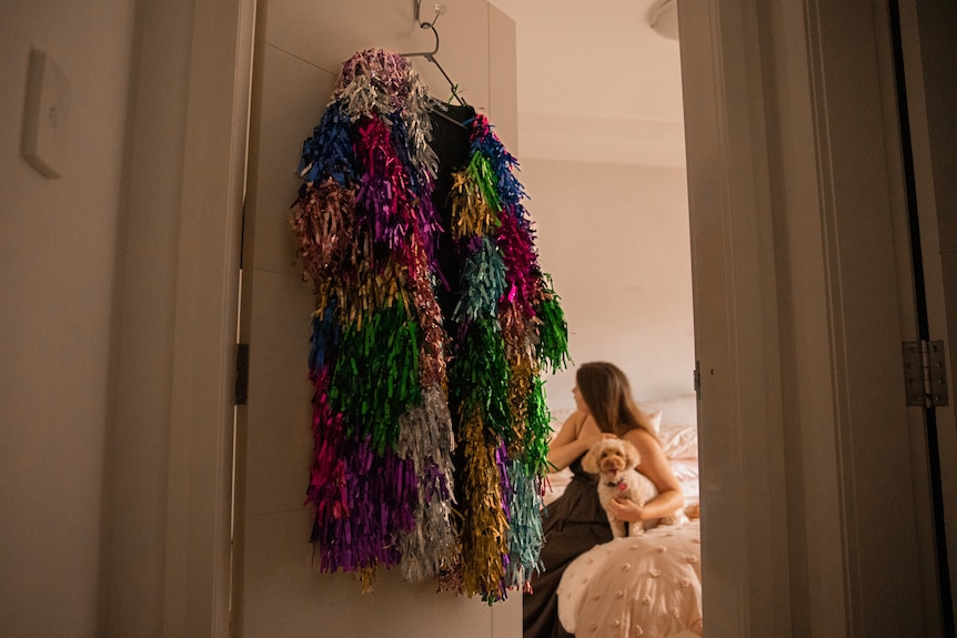 A woman sits on her bed brushing her hair with her dog. A rainbow tinsel jacket hangs off the door.