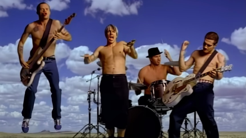 Four shirtless members of Red Hot Chili Peppers playing instruments in a green field before a blue sky.
