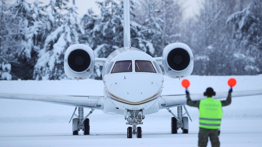 A private jet aircraft on a snowy airstrip, being directed by a man in high-viz holding light padels.