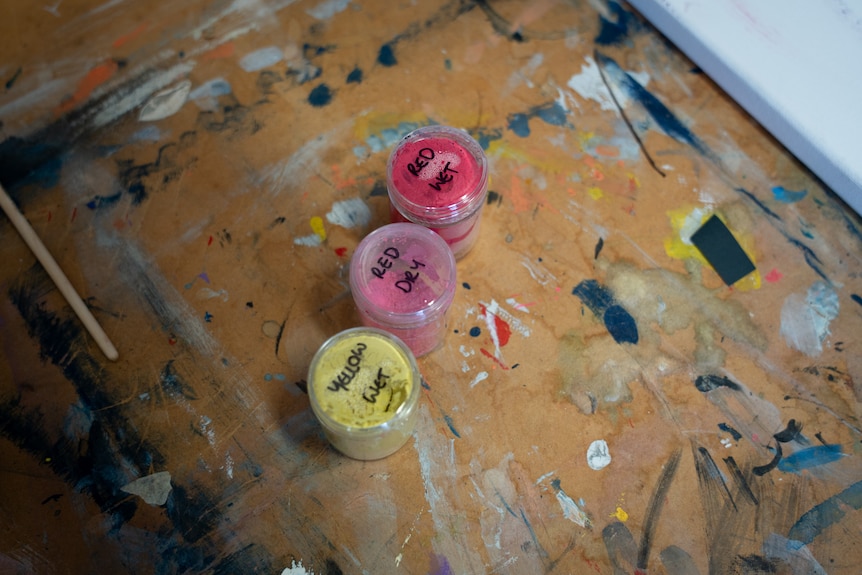 俯视图，克里沾满油漆的木桌上放着三罐粉色、红色和黄色的干燥颜料。 
