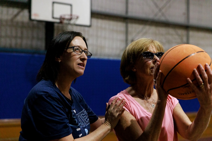 A women wearing glasses has her arms placed on an older woman's shoulders to help her as she prepares to shoot the basketball