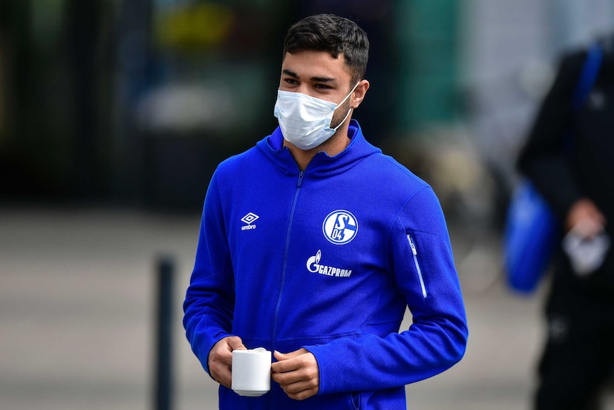 Ozan Kabak with a Schalke tracksuit top, holding a cup and wearing a facemask.