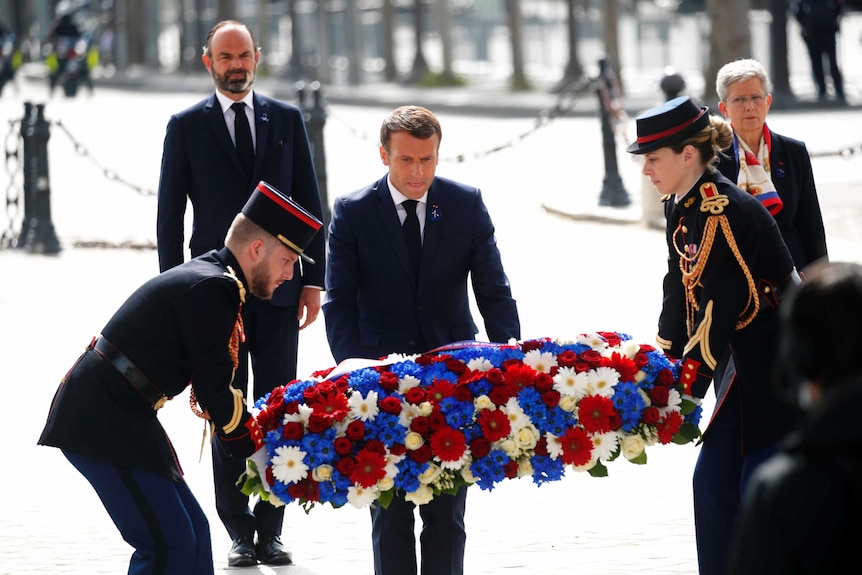 a man in a suit bends down to lay a wreath, assisted by two people in uniform.