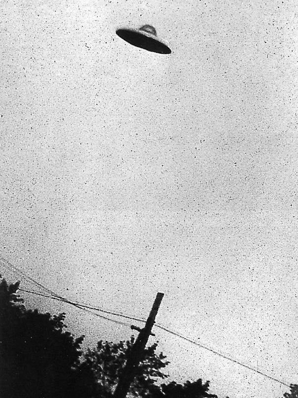 A grainy black and white photograph shows a disc-shaped object hovering over a suburban street.
