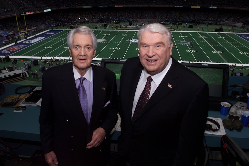 Dos locutores, el de la derecha, un ex entrenador de la NFL, aparecen en una cabina con vista al campo antes del Super Bowl.