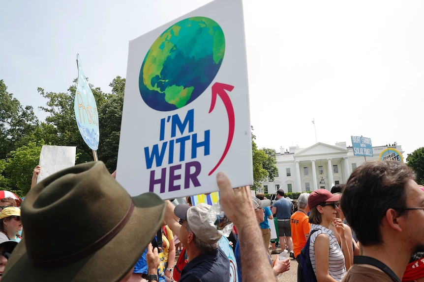 A man holds a sign reading "I'm with her" which points to an illustration of the eart, standing outside the white house