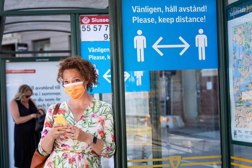 A woman in a floral dress and face mask waits at a bus stop