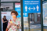A woman in a floral dress and face mask waits at a bus stop