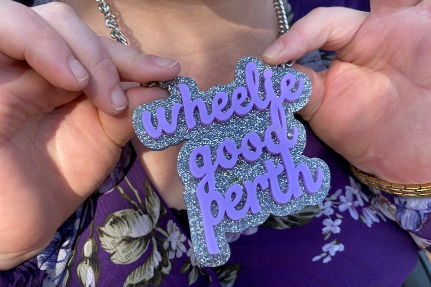 A close up shot of a graphic purple necklace which reads 'Wheelie Good Perth'