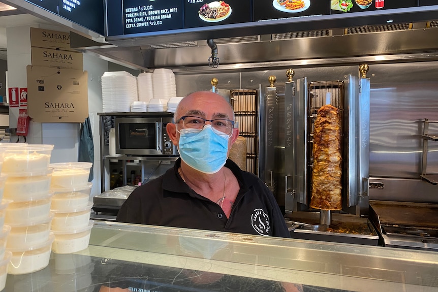 A man wearing a mask stands behind a counter and in front of a kebab rotisserie.