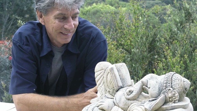 A man smiles at a stone statue at a green lookout.