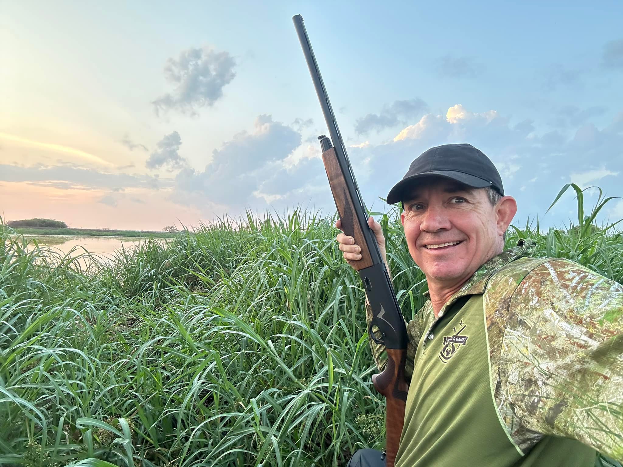 Um homem com uma camisa camuflada tira uma selfie com uma arma em pé na grama alta perto do lago