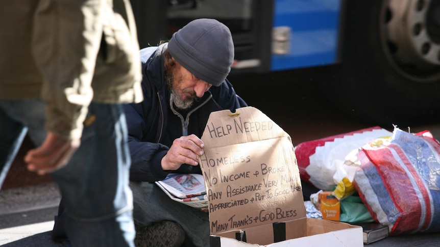 A man begging on the street with a cardboard sign as people are walking by him.