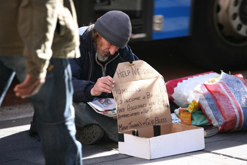 A man begging on the street with a cardboard sign as people are walking by him.