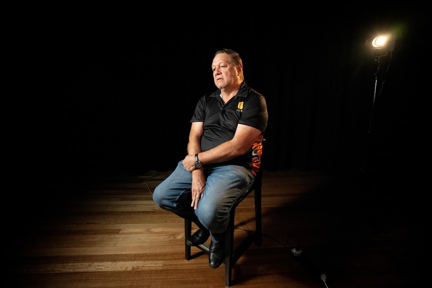 A man sits in a dark room being interviewed.