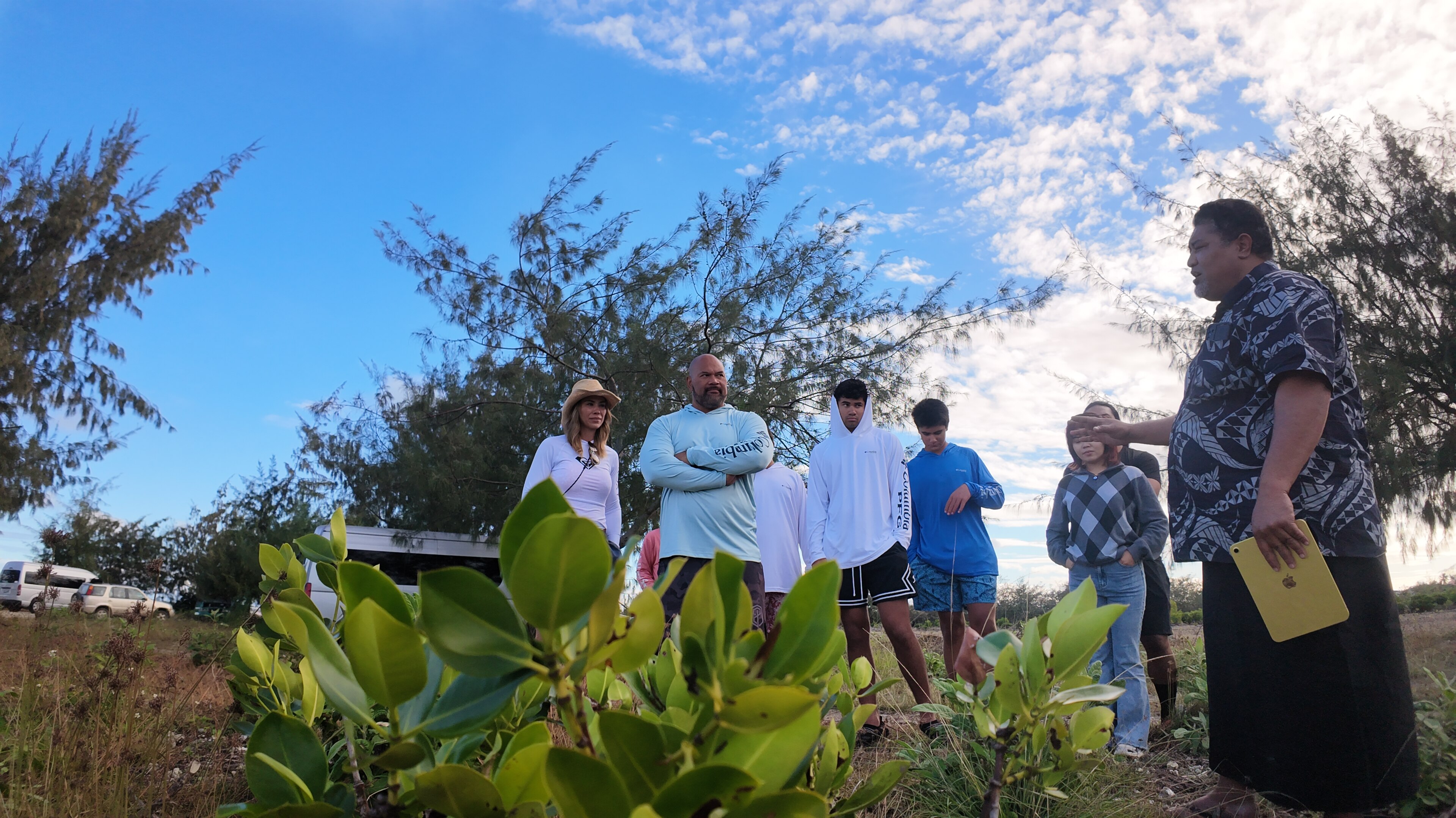 A Tongan man and his family stand behind some green plants and listen to a man standing to the side with his arm out explaining