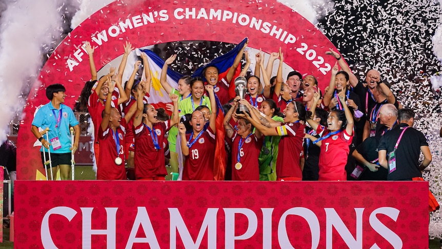 Members of the Philippines women's football team hold a trophy aloft and celebrate with arms raised.