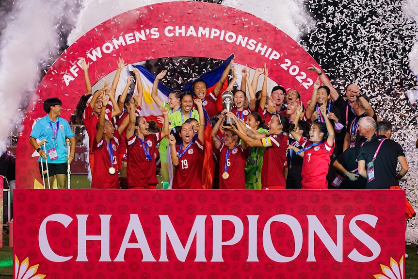 Members of the Philippines women's football team hold a trophy aloft and celebrate with arms raised.