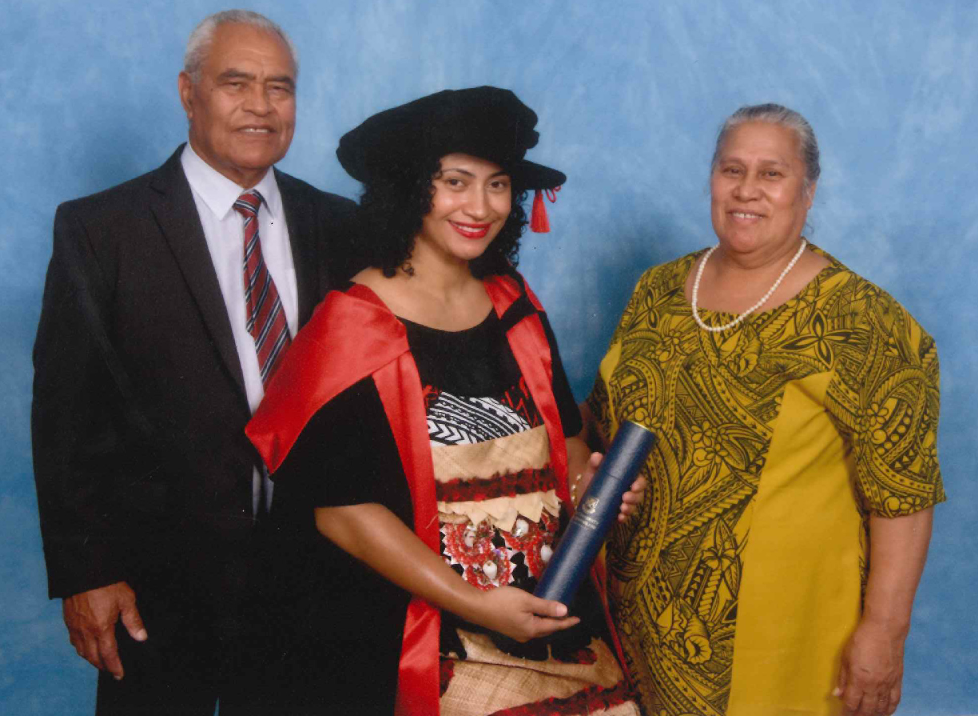 Woman wears traditional Tongan attire under her black graduation gown, with parents by her side smiling.
