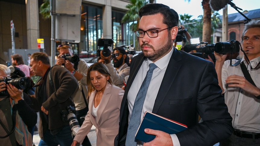 A man walks out of glass doors flanked by a lawyer and journalists.