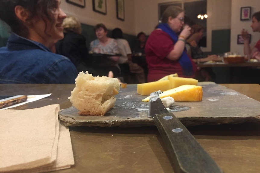A close up of a chunk of bread and wedges of cheese on a slate cheeseboard with diners in the background.