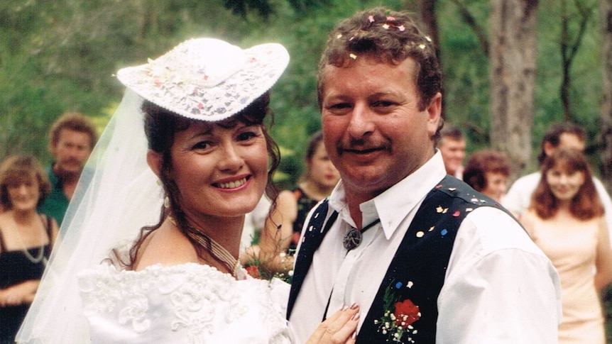 Wedding photo with a woman in a white dress and hat and man in a suit, with people in the background.