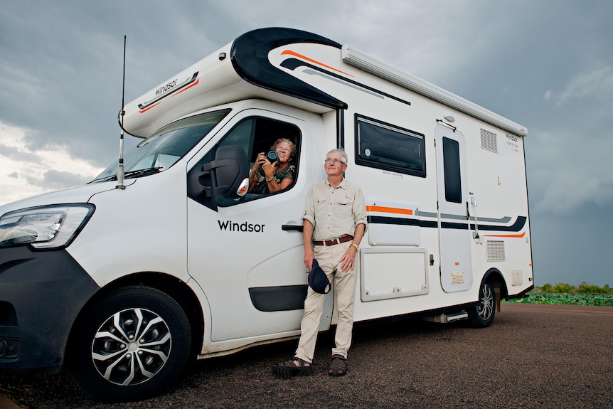 A motorhome with a woman in the front seat, holding a camera, and a man standing next to the door. 