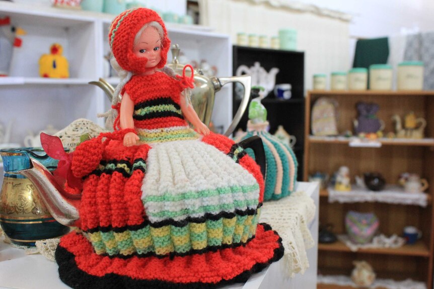 Red knitted rag doll tea cosy on shelf.