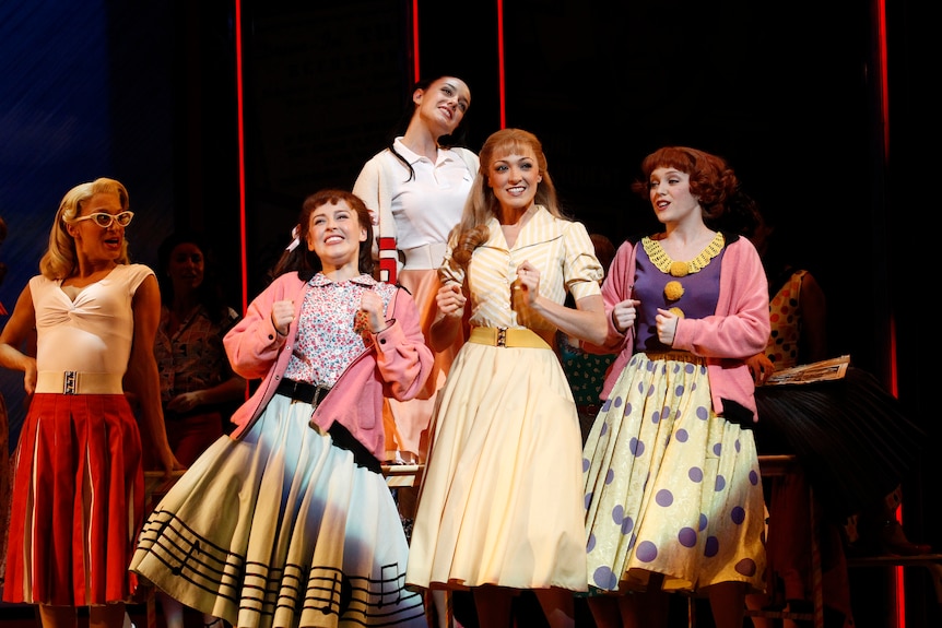 A group of five women dressed in 1950s clothing, performing on stage. 