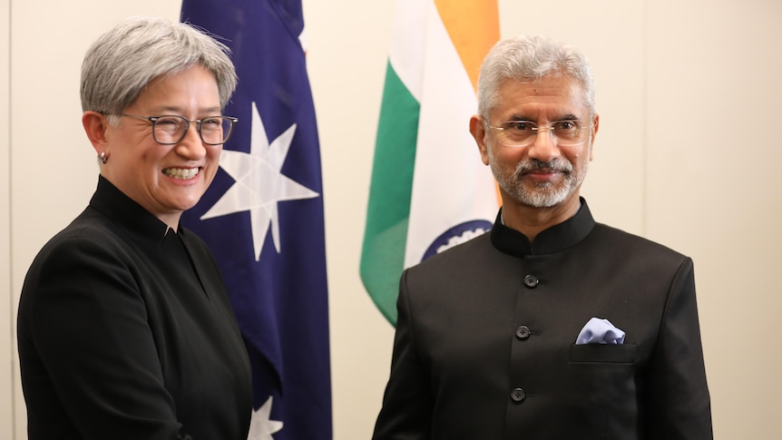 A woman and a man stand in front of the Australian and Indian flags shaking hands. 