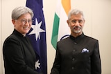 A woman and a man stand in front of the Australian and Indian flags shaking hands. 