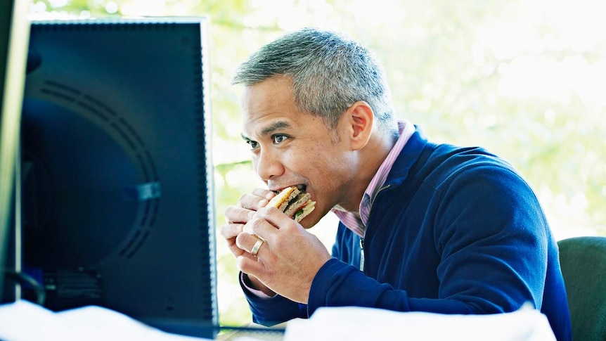 A businessman eats lunch at a desk in his office while watching a computer screen.