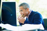 A businessman eats lunch at a desk in his office while watching a computer screen.