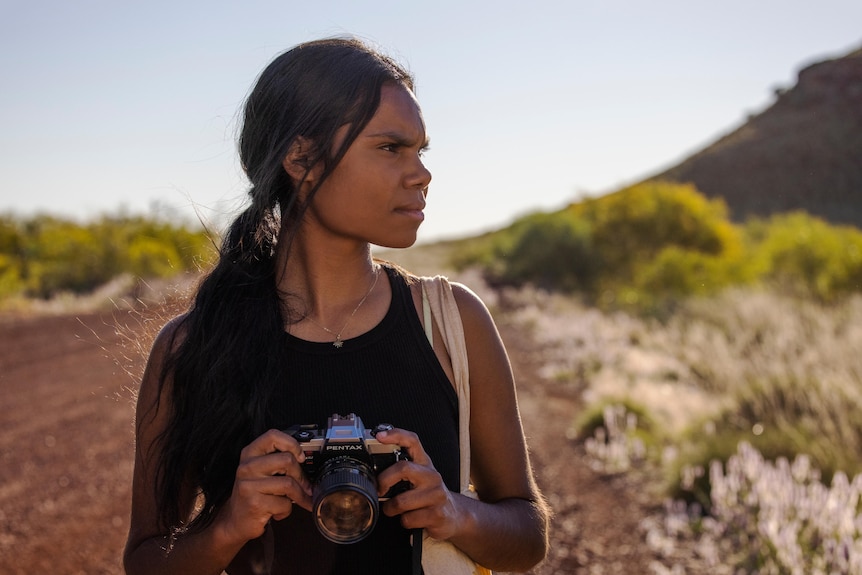 A young Indigenous woman stands on a red dirt road in the outback, holding a camera to her chest.