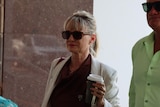 A woman in a blazer and wearing sunglasses walks alongside a man also wearing sunglasses