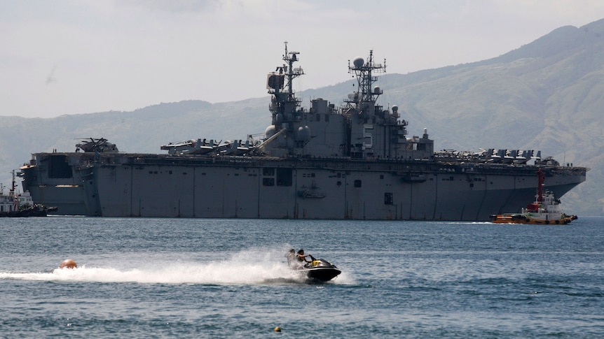 A ship is seen in the water with two people on a jet ski in front. Mountainous land is seen behind, and the air is hazy.