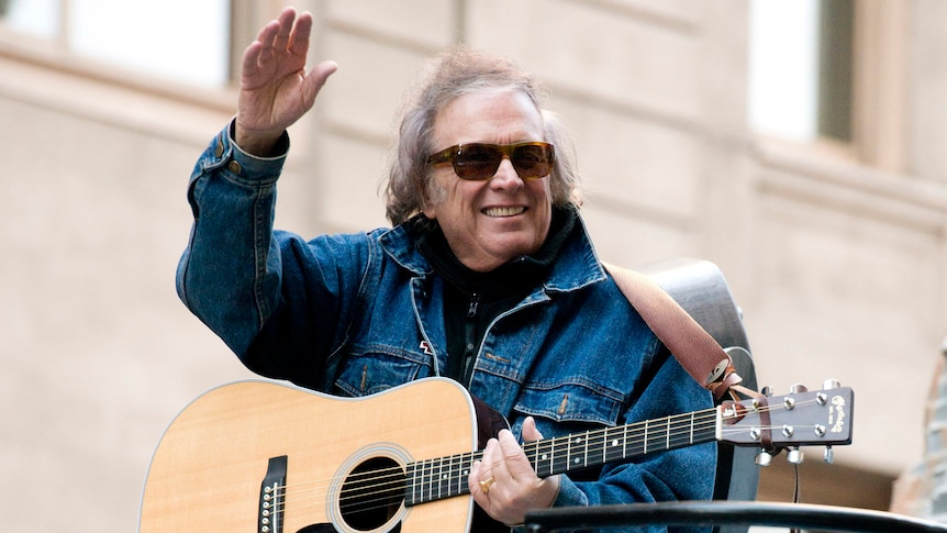 Don McLean, wearing shades and a denim jacket and carrying an acoustic guitar, waves to a crowd during a thanksgiving parade